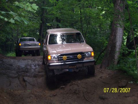 1979 Ford F-150 - Two Ford Broncos navigate a challenging off-road path through a wooded area.