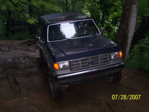 1979 Ford F-150 - A dark blue Ford Bronco navigates a challenging muddy trail through a wooded area.