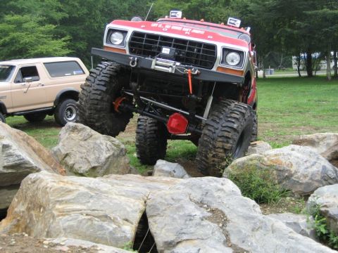 1995 Ford Bronco - A heavily modified red Ford Bronco navigates a rocky obstacle course, with another Bronco parked nearby.