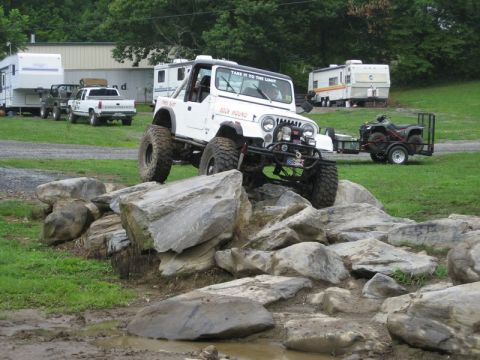 1995 Ford Bronco - A modified white Jeep Wrangler navigates a rocky obstacle course, with other vehicles and campers in the background.