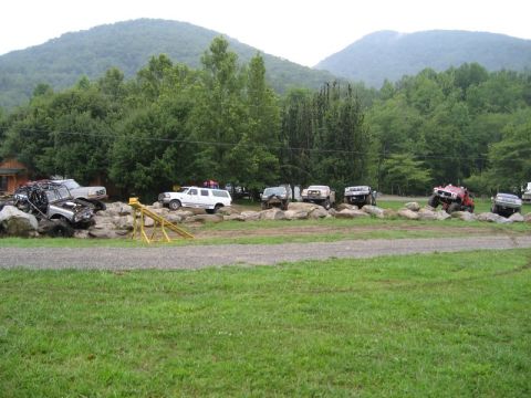 1995 Ford Bronco - Multiple off-road vehicles traverse a challenging rocky terrain, set against a backdrop of lush trees and rolling hills.