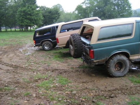 1995 Ford Bronco - Three Ford Broncos are parked on a muddy, grassy area with trees in the background.