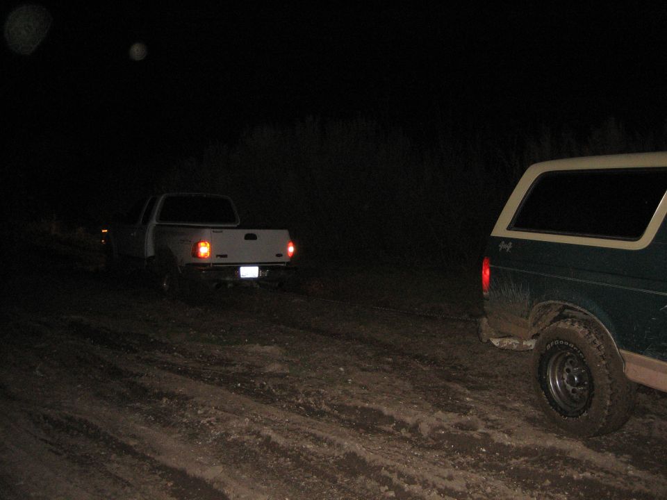 Green SUV and white pickup truck on a dirt road at night. The SUV is in the foreground, right side. The pickup is in the background, left side