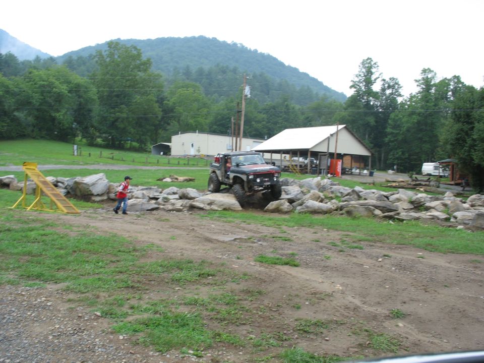 Off-road vehicle with large tires driving over rocks. A person in a red shirt walks nearby. Buildings and trees are in the background