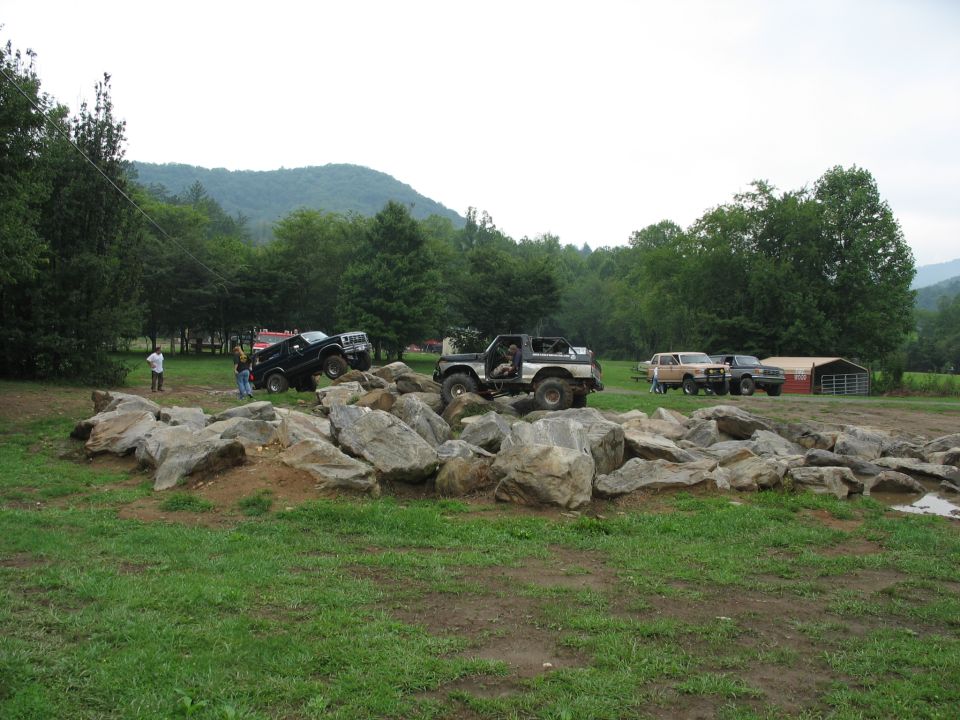 Off-road vehicles on a rock obstacle course with spectators and a barn in the background
