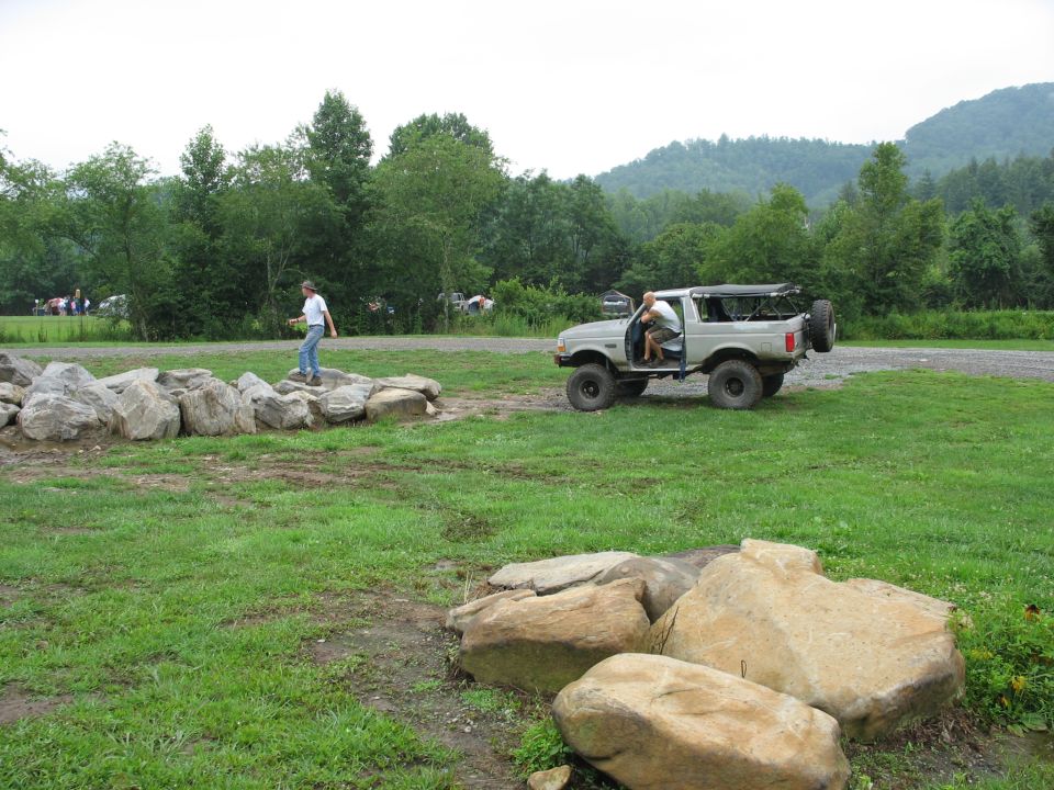 Gray Ford Bronco with soft top and spare tire on rear, driver's door open, parked on dirt and grass