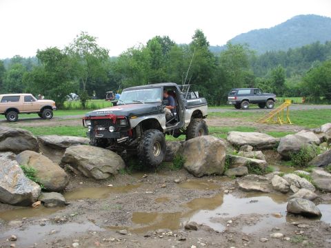1995 Ford C-350 (Centurion Conversions) - A heavily modified pickup truck with large tires maneuvers through a rocky obstacle course, with other vehicles visible in the background.