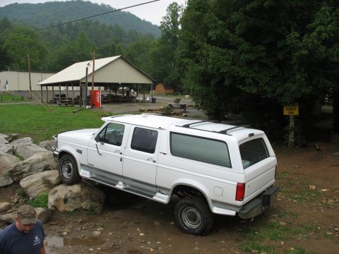 1995 Ford C-350 (Centurion Conversions) - A white extended SUV navigates a rocky, muddy terrain with a covered picnic area and trees in the background.