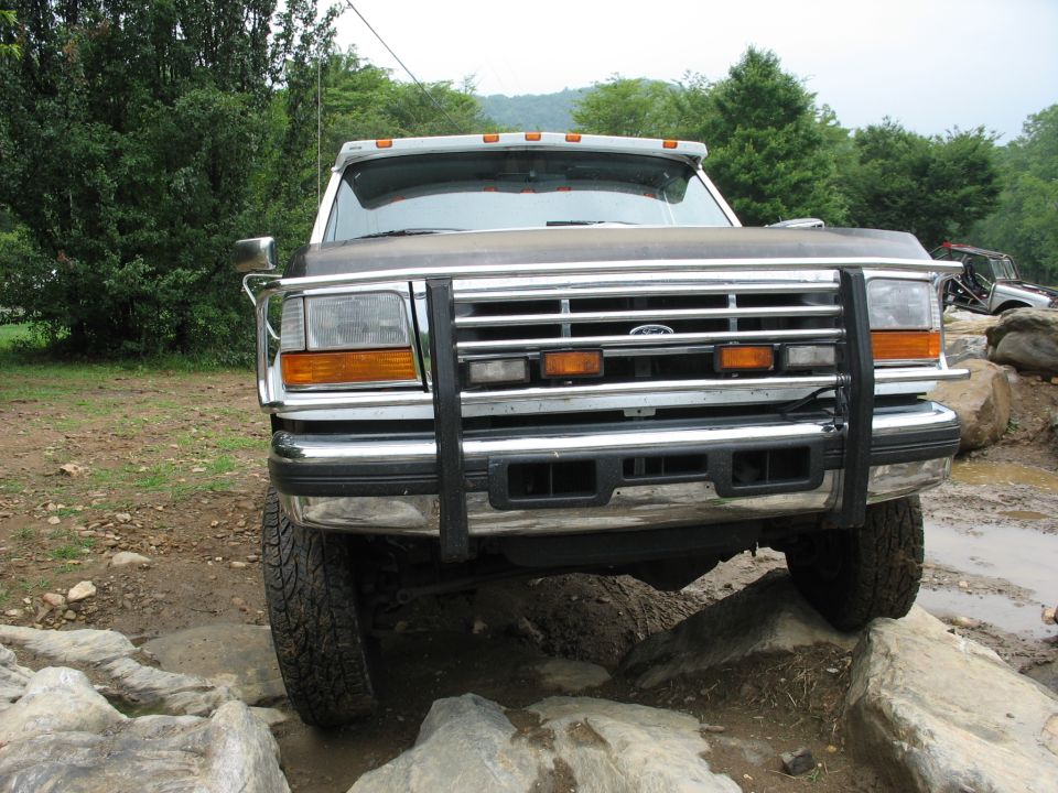 Front view of a dark-colored Ford F-Series truck with a chrome grille guard, navigating rocky terrain