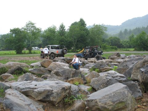 1995 Ford C-350 (Centurion Conversions) - People and off-road vehicles are gathered in a rocky outdoor area with trees and hills in the background.