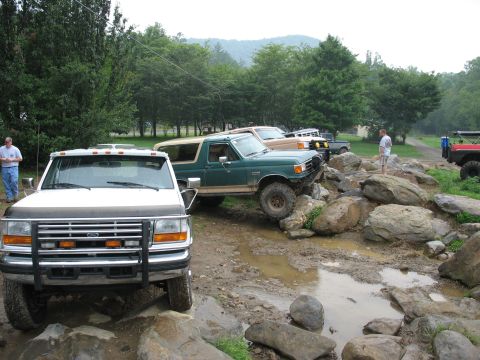 1995 Ford C-350 (Centurion Conversions) - A white 1995 Ford F-Series truck is parked in the foreground while other vehicles navigate a rocky, muddy off-road course.