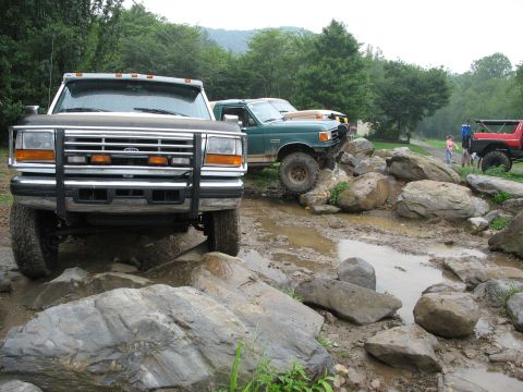 1995 Ford C-350 (Centurion Conversions) - Several trucks traverse a challenging off-road course filled with large rocks and mud puddles.
