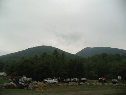 1995 Ford C-350 (Centurion Conversions) - A collection of off-road vehicles are parked on a rocky area, with a dense forest and mountains visible behind them.
