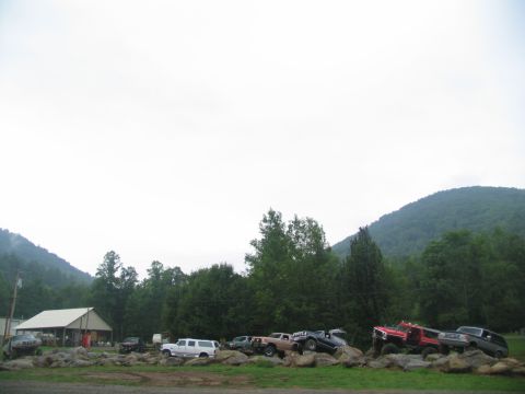 1995 Ford C-350 (Centurion Conversions) - Multiple off-road vehicles are parked on a rocky terrain in front of a building and wooded hills.