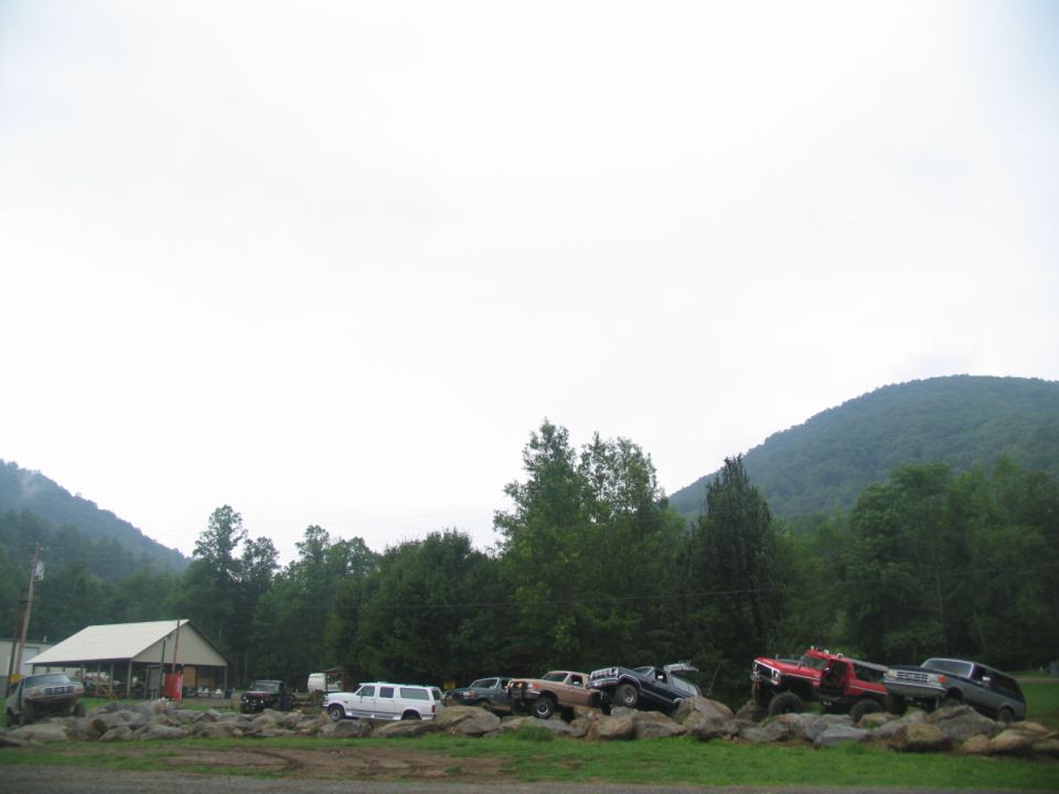 Several modified off-road trucks and SUVs parked on large rocks with a building and trees in the background