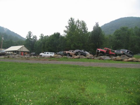 1995 Ford C-350 (Centurion Conversions) - Multiple off-road vehicles are positioned on a rocky terrain, with a building and wooded hills in the distance.