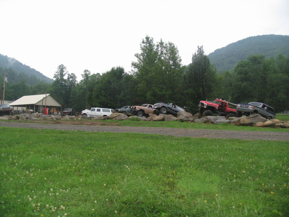 Trucks and SUVs parked atop a rock pile beside a rural building and forested mountains