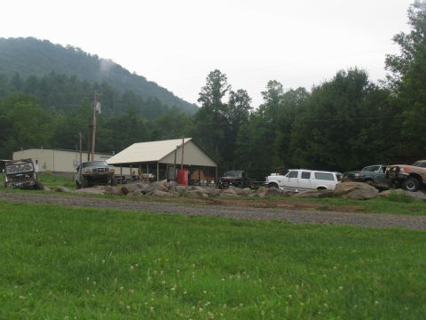 1995 Ford C-350 (Centurion Conversions) - A white SUV is among several vehicles parked in a lot near a covered structure and a wooded hillside.