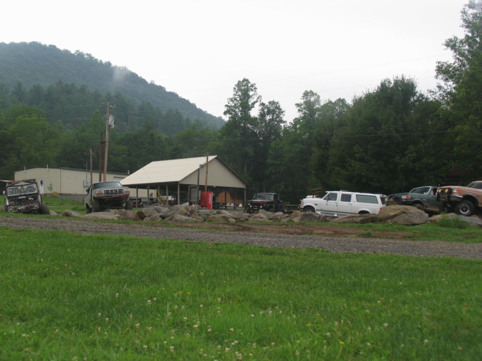 White SUV parked near a building with other vehicles and a wooded hill in the background