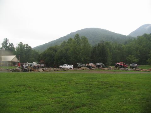 1995 Ford C-350 (Centurion Conversions) - Multiple off-road vehicles are positioned on a rocky terrain, with a forested mountain range visible behind them.