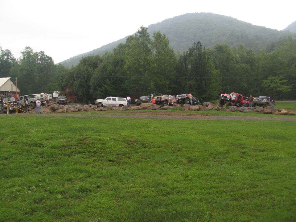Several off-road trucks and SUVs are positioned on a rocky terrain with a wooded hillside in the background