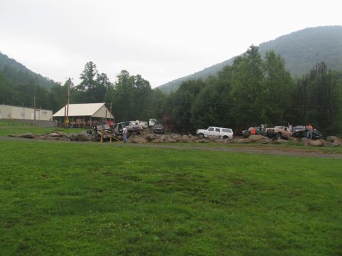 1995 Ford C-350 (Centurion Conversions) - Several trucks and SUVs are gathered on a rocky terrain with a building and wooded hills in the background.