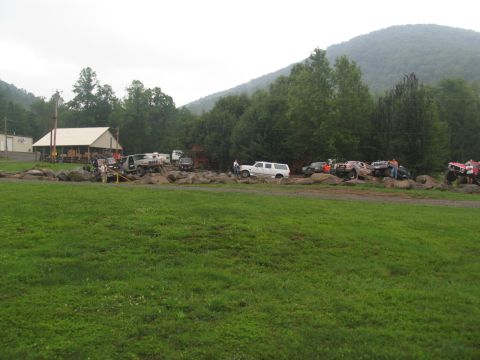 1995 Ford C-350 (Centurion Conversions) - Several off-road vehicles are parked on a rocky area in front of a building and trees.