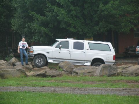 1995 Ford C-350 (Centurion Conversions) - A woman stands beside a white extended SUV that is parked on a rocky surface near a building.