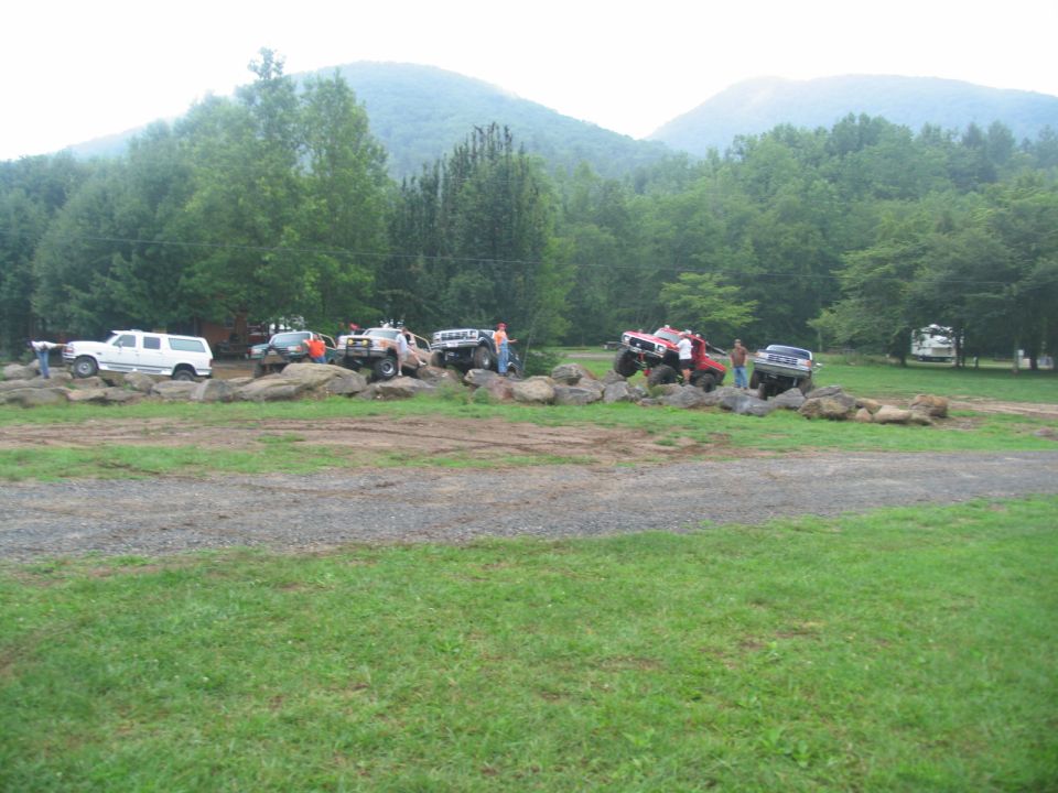 Multiple off-road vehicles maneuvering over a rocky obstacle course with trees and mountains in the background
