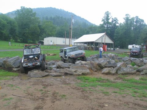 1995 Ford C-350 (Centurion Conversions) - Off-road vehicles tackle a rocky obstacle course with a mountainous backdrop and buildings in the distance.