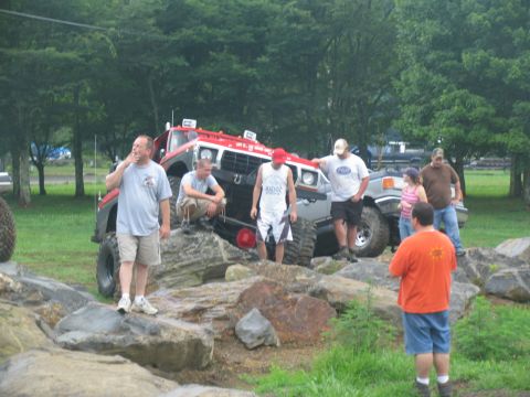 1995 Ford C-350 (Centurion Conversions) - Several people stand around modified off-road trucks navigating a rocky terrain.