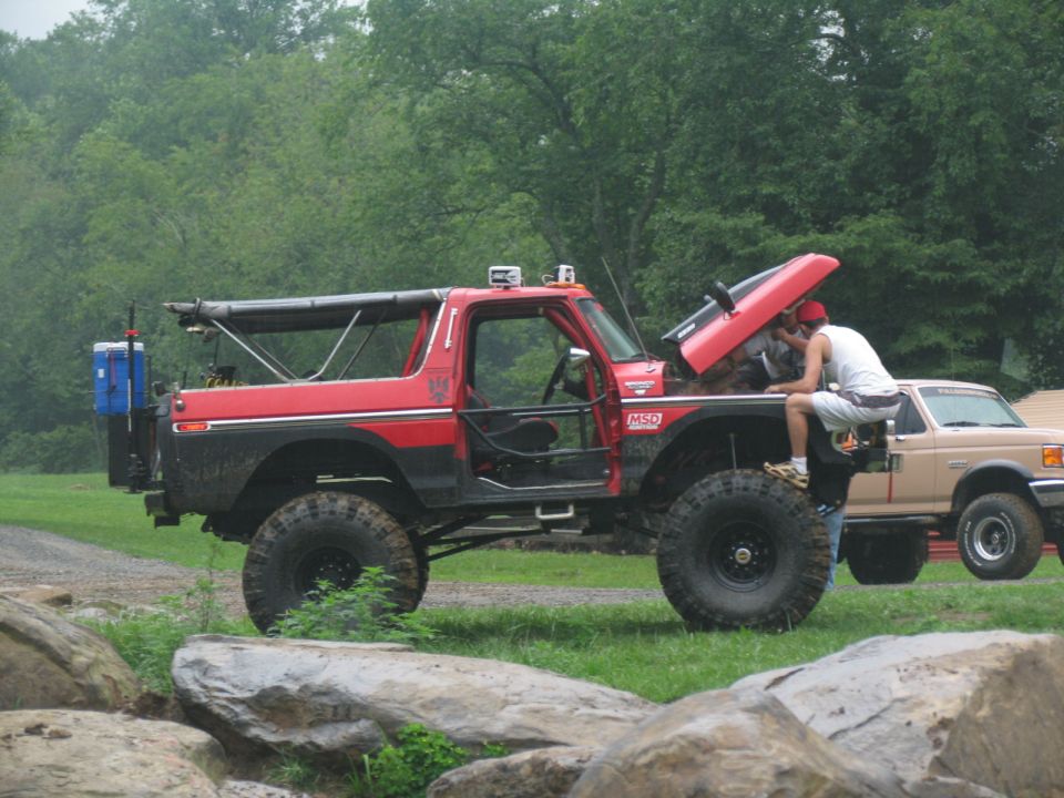 Red off-road SUV with hood open, person working on engine. Another tan truck is visible behind it
