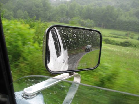 1995 Ford C-350 (Centurion Conversions) - A white vehicle and a dark SUV drive on a wet road surrounded by lush green scenery.