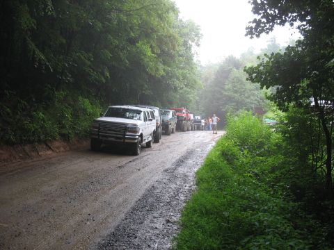 1995 Ford C-350 (Centurion Conversions) - A line of trucks and SUVs are parked on a wet, gravel road surrounded by trees.