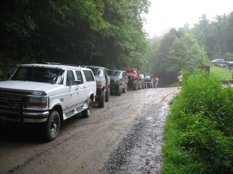 1995 Ford C-350 (Centurion Conversions) - A convoy of modified trucks and SUVs, including a white Ford conversion, navigate a wet dirt road through a wooded area.