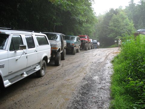 1995 Ford C-350 (Centurion Conversions) - Several off-road vehicles are lined up on a muddy, gravel road in a wooded area.