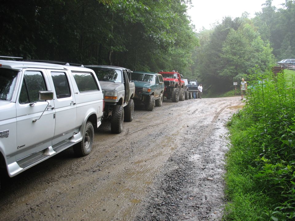 Line of modified trucks and SUVs parked on a wet dirt road with trees on either side