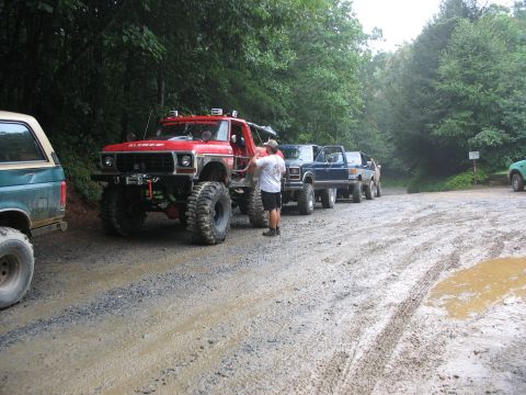 1995 Ford C-350 (Centurion Conversions) - Several off-road vehicles are parked on a wet, gravelly surface surrounded by trees.