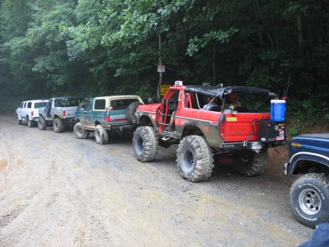 1995 Ford C-350 (Centurion Conversions) - A line of off-road vehicles, including a red Bronco, are parked on a dirt road in a wooded area.