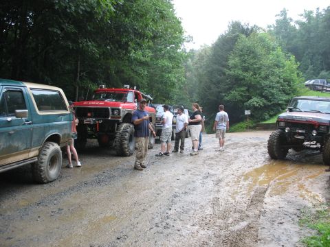1995 Ford C-350 (Centurion Conversions) - A group of people gather around modified off-road vehicles on a wet, muddy track surrounded by trees.