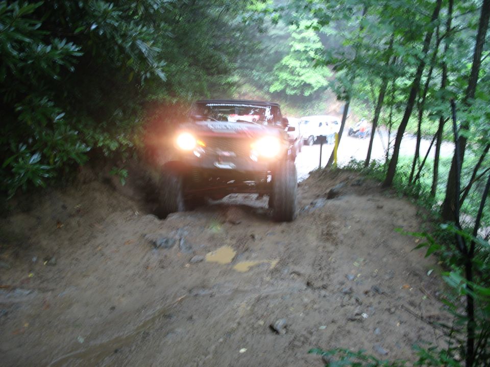 Off-road vehicle driving through mud on a wooded trail, headlights on