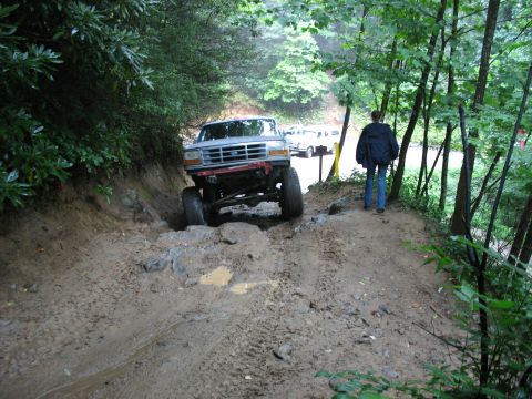 1995 Ford C-350 (Centurion Conversions) - A lifted Ford F-Series truck navigates a challenging muddy terrain with trees on either side.