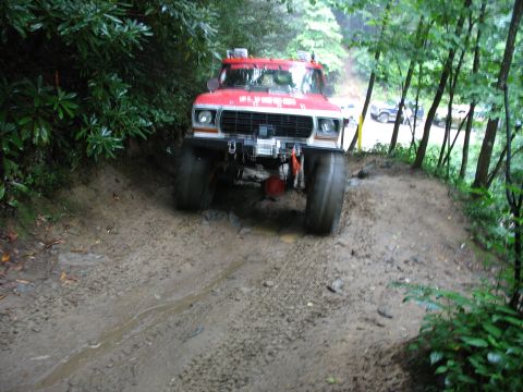 1995 Ford C-350 (Centurion Conversions) - A red truck navigates a wet, muddy path through a wooded area, with other vehicles visible in the background.