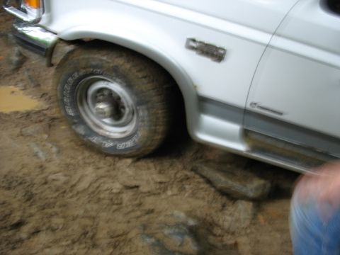 1995 Ford C-350 (Centurion Conversions) - A white pickup truck navigates through a muddy and rocky terrain, with its front tire partially submerged.