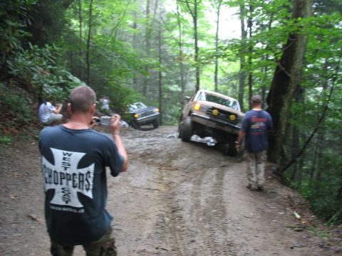 1995 Ford C-350 (Centurion Conversions) - Two off-road vehicles navigate a challenging muddy path in a dense forest, with onlookers documenting the action.