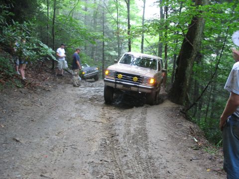 1995 Ford C-350 (Centurion Conversions) - Two trucks navigate a challenging muddy trail in a wooded area.