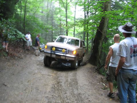 1995 Ford C-350 (Centurion Conversions) - A group of people watch as a large SUV navigates a difficult, muddy off-road path in a wooded area.
