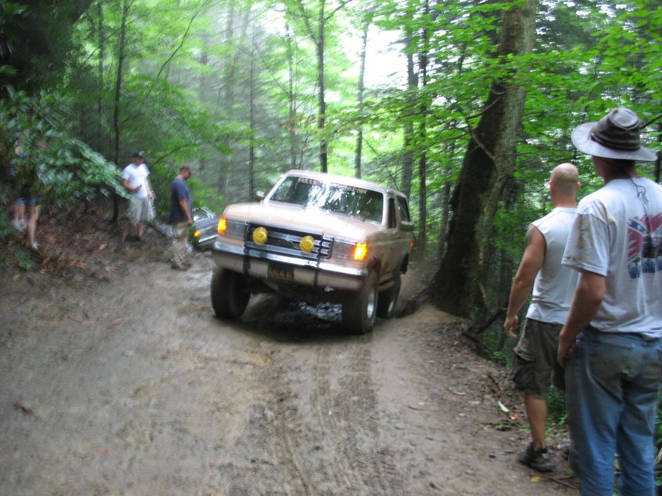 Brown SUV driving on a muddy forest trail with people watching