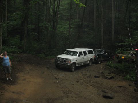 1995 Ford C-350 (Centurion Conversions) - A white 1995 Ford C-350 Centurion Conversions SUV navigates a muddy path through a dense forest, with other vehicles following behind.
