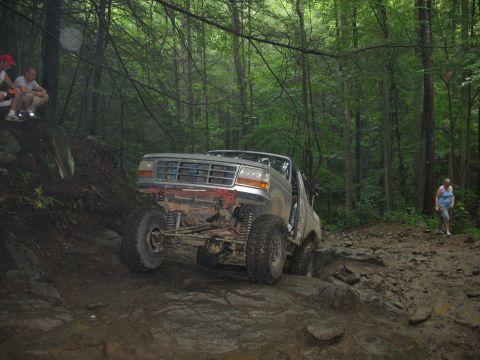 1995 Ford C-350 (Centurion Conversions) - Off-road truck with modified suspension tackles a challenging, wet, and rocky path surrounded by dense trees.